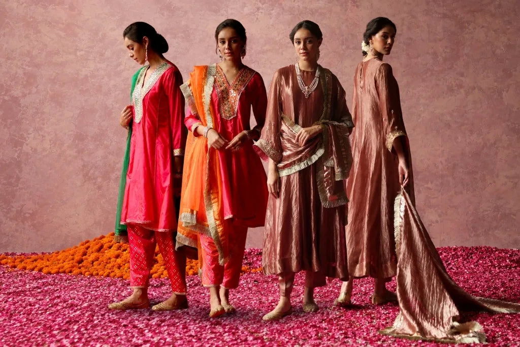 Four women wearing colorful ethnic outfits with embroidered kurtas, pants, and dupattas.